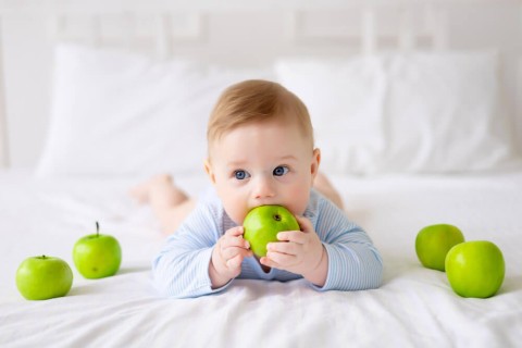 cute-healthy-little-baby-is-lying-bed-white-bedding-home-blue-bodysuit-baby-holds-his-hands-tastes-green-apples-first-complementary-food-healthy-food-1
